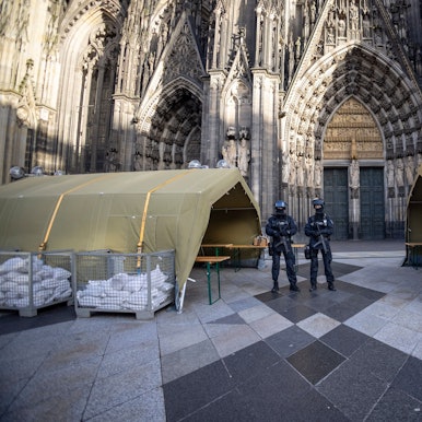 Polizisten stehen mit Maschinenpistolen vor dem Kölner Dom. Neben ihnen stehen Zelte.