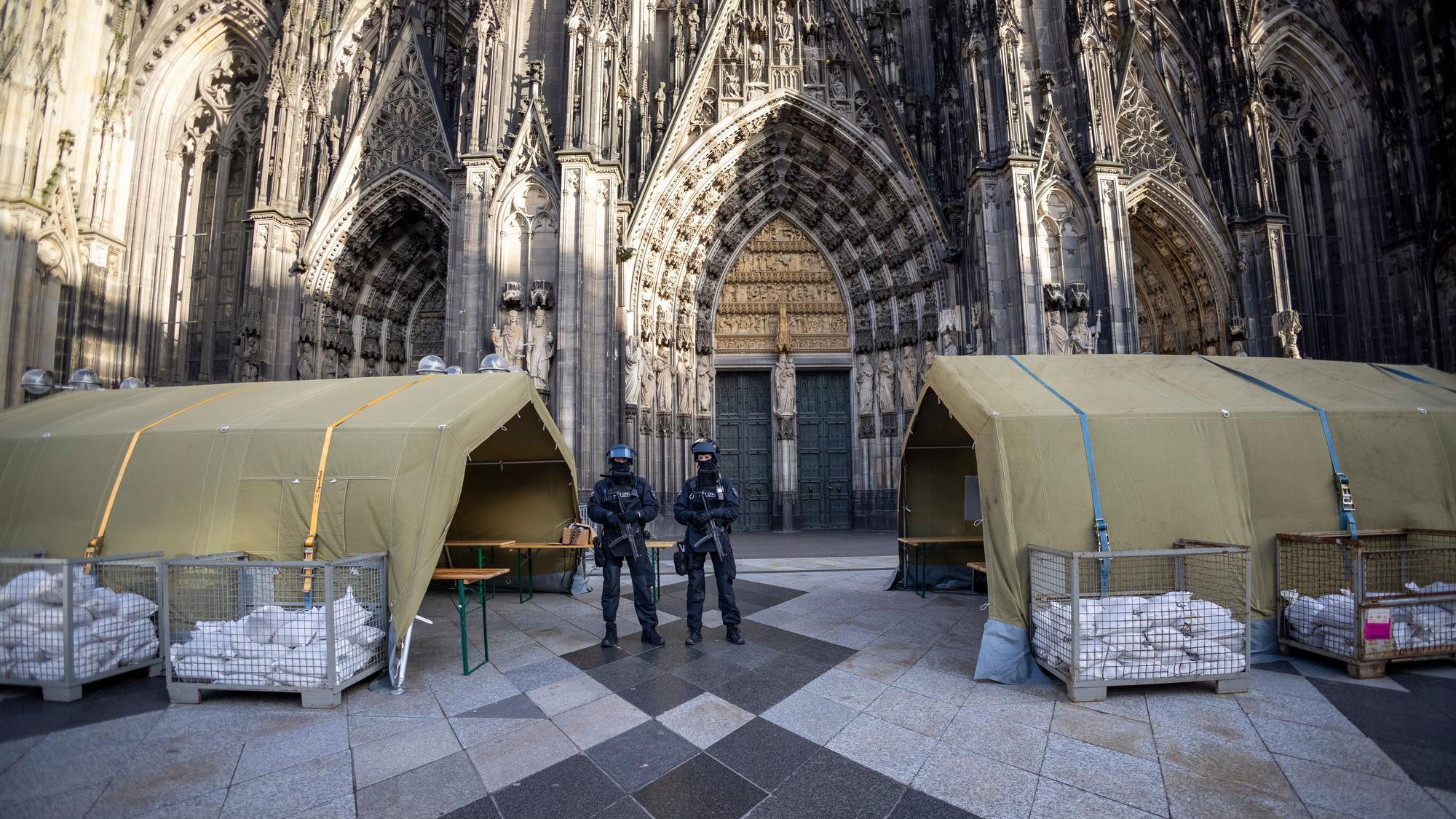 Polizisten stehen mit Maschinenpistolen vor dem Kölner Dom. Neben ihnen stehen Zelte.