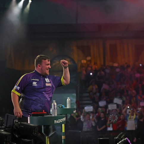Der Jugendliche Dartspieler Luke Littler hebt triumphierend die Faust vor einer Menge.