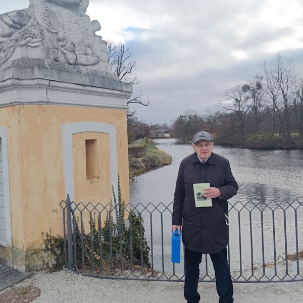 Ein älterer Mann hält ein Buch in die Kamera, hinter ihm ist eines der Gewässer im Park von Schloss Augustusburg zu sehen.