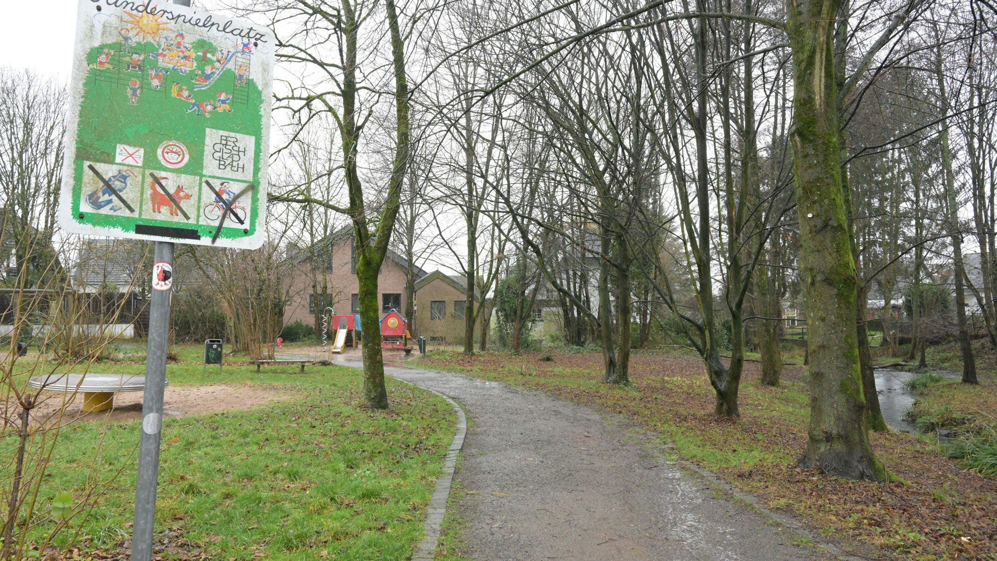 Im Vordergrund ist ein Spielplatz-Schild. Links neben dem Weg sieht man Spielgeräte, rechts neben dem Weg stehen Bäume.