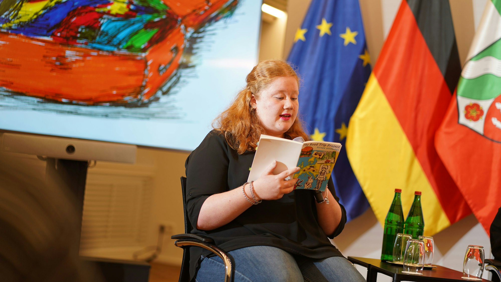 Marie Emonds liest vor. Hinter ihr stehen die Flaggen der Bundesrepublik Deutschland, von NRW und Europa..