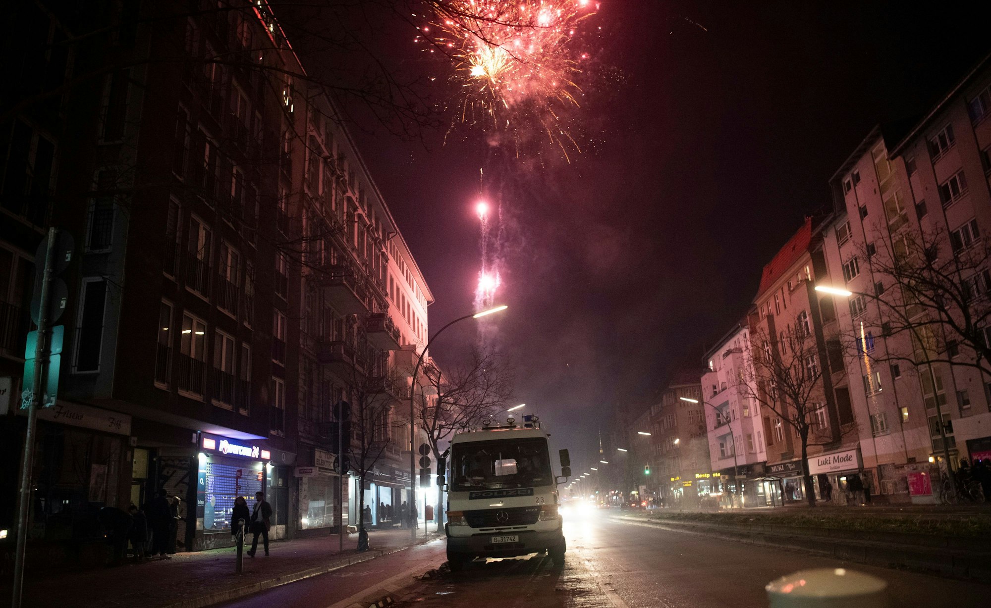 Ein größeres Polizeifahrzeug steht im Dunkeln am Straßenrand zwischen Häusern. Im Hintergrund explodieren Feuerwerkskörper am Nachthimmel.