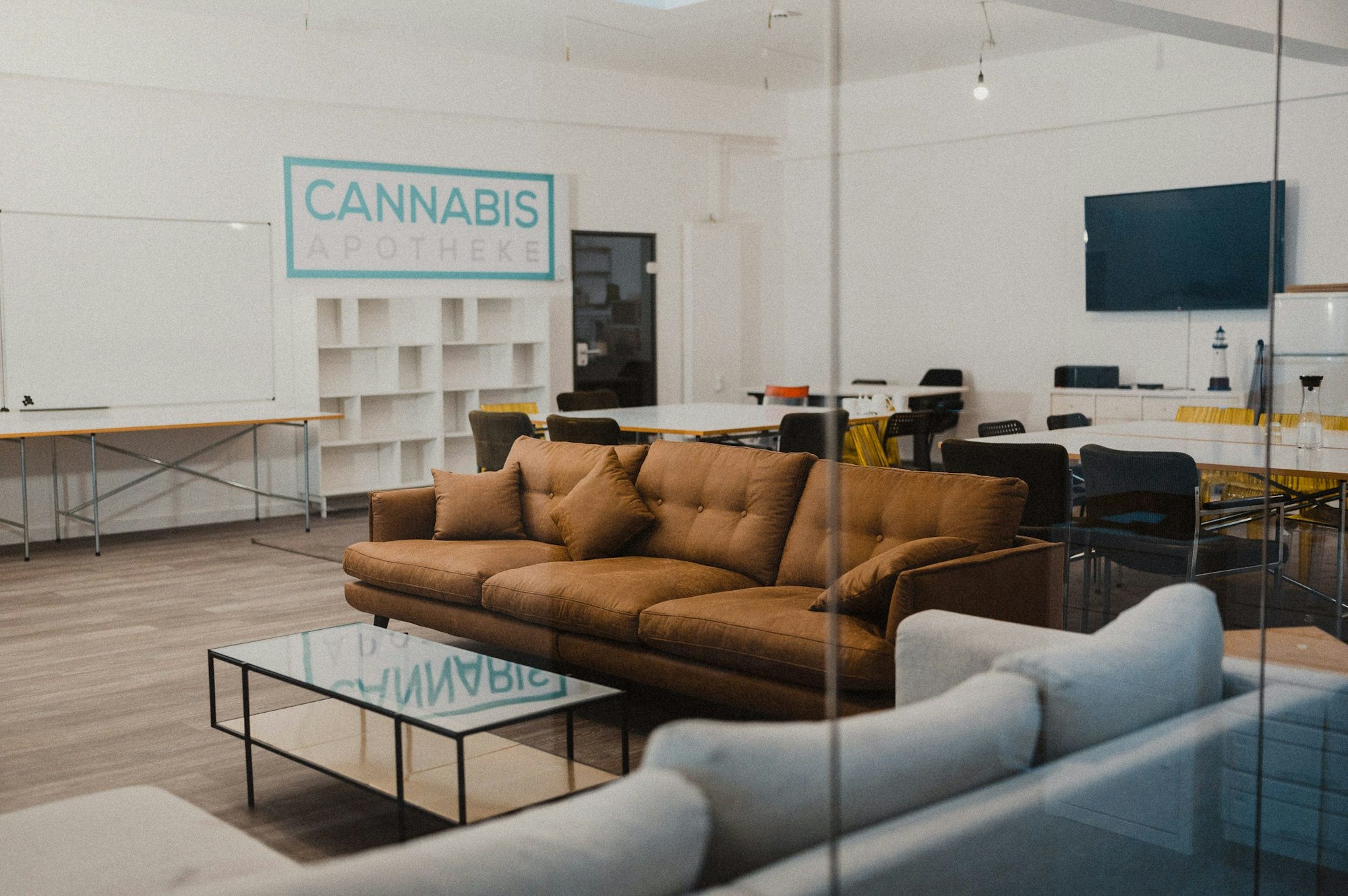 Ein Sofa in einem Seminarraum mit Tischen und Regalen im Hintergrund. Auf der Wand steht der Schriftzug “Cannabis Apotheke”.