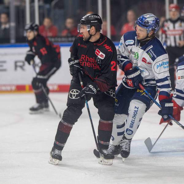 Zwei Eishockeyspieler stehen nah beieinander vor einem Tor mit einem weiteren Spieler.