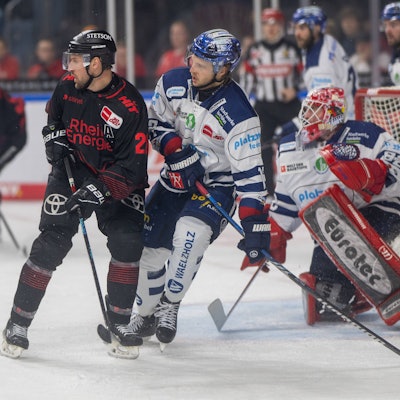 Zwei Eishockeyspieler stehen nah beieinander vor einem Tor mit einem weiteren Spieler.