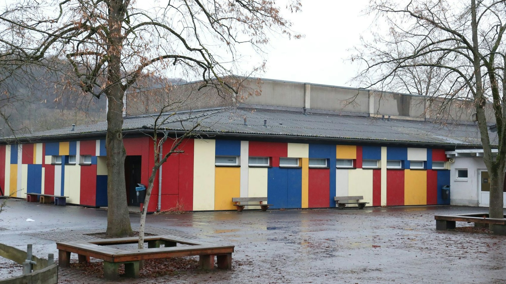 Vor einem flachen Gebäude mit einer bunten Fassade stehen zwei Bäume und einige Sitzbänke.