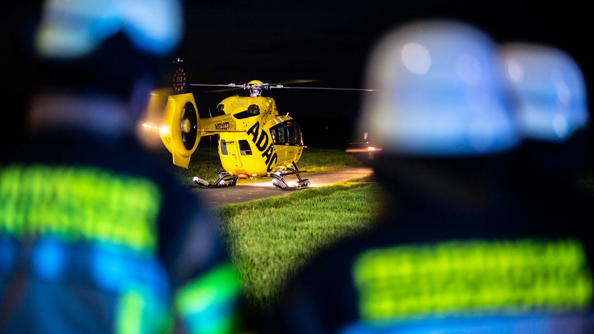 Das Bild zeigt einen Rettungshubschrauber, der auf einem Wirtschaftsweg in der Nacht gelandet ist. Drei Feuerwehrleute sind im Vordergrund zu sehen.