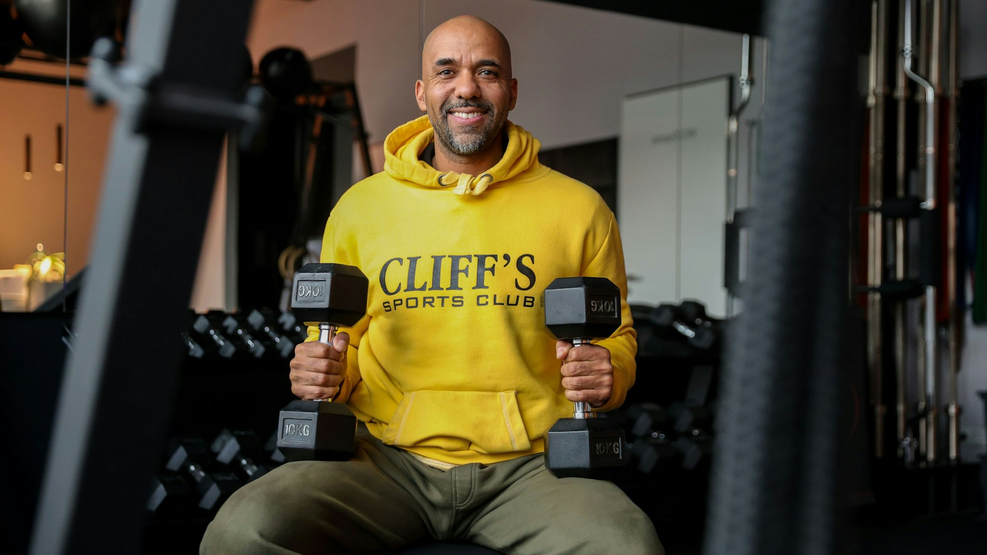Clifford Opoku-Afari sitzt mit Hanteln in der Hand in einem Sportstudio.