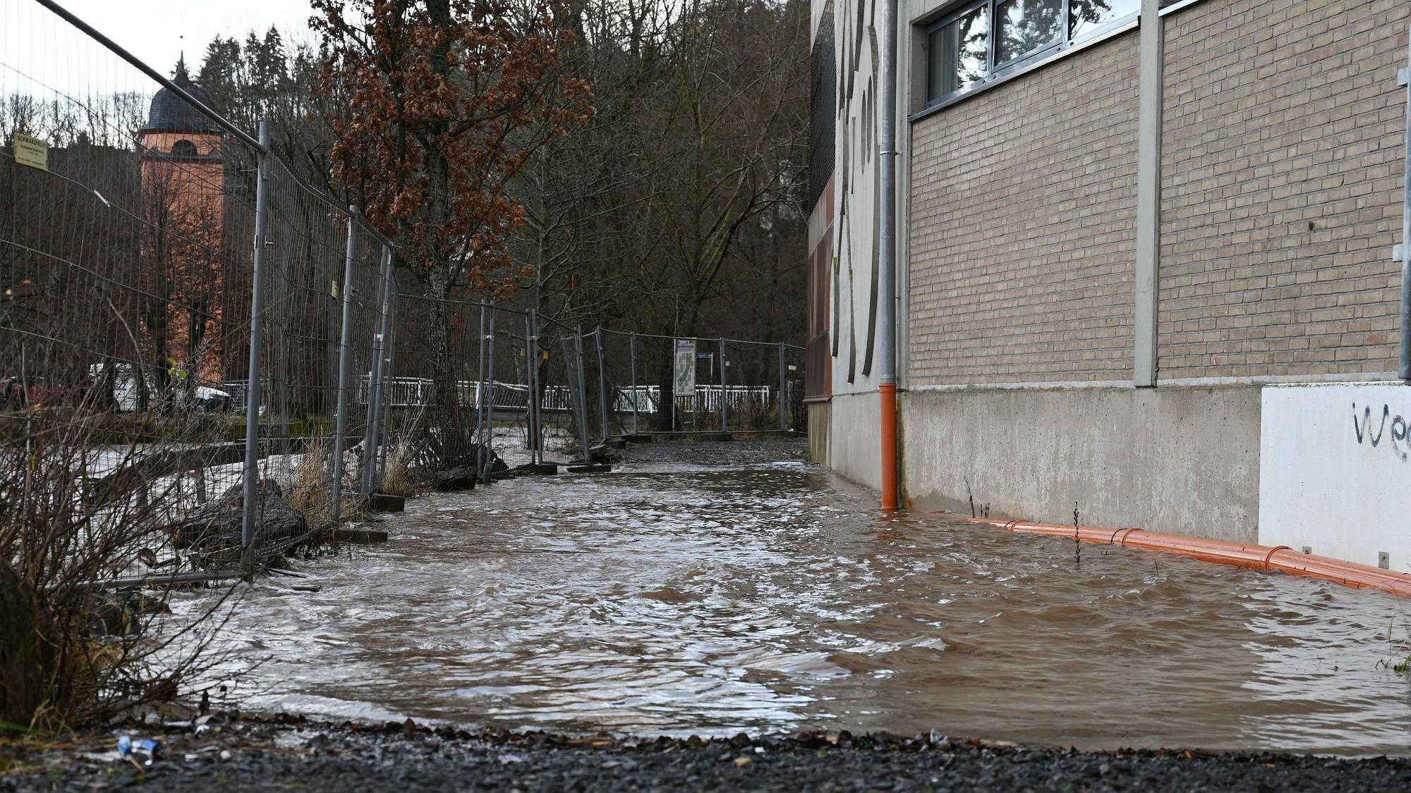 Links hinter einem Bauzaun ist die Olef in Schleiden. Der Fluss führt Hochwasser, das Wasser reicht bis ans Sturmius-Gymnasium heran.