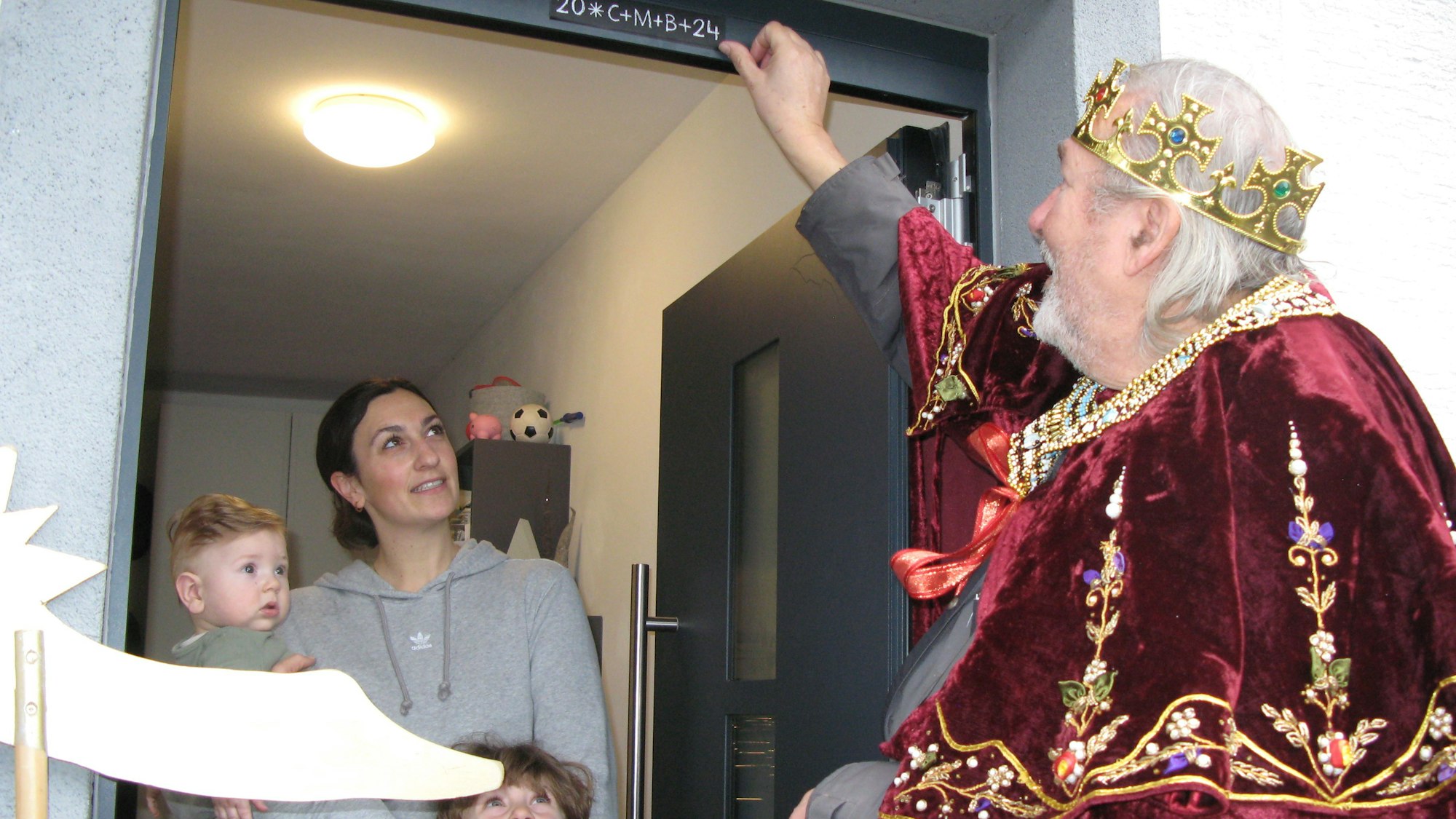 Sternsinger Wolfgang Czarnecki bringt über einer geöffneten Haustür den Segensaufkleber an, eine Frau mit einem kleinen Jungen und einem Baby auf dem Arm steht in der Tür und schaut zu.