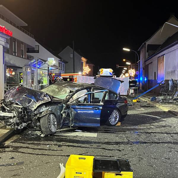 Ein zertrümmertes, dunkles Auto steht quer auf einer Straße, im Hintergrund ist das verbogene Gestänge des Wartehäuschens zu sehen.
