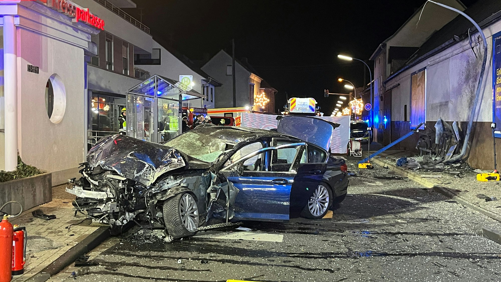 Ein zertrümmertes, dunkles Auto steht quer auf einer Straße, im Hintergrund ist das verbogene Gestänge des Wartehäuschens zu sehen.
