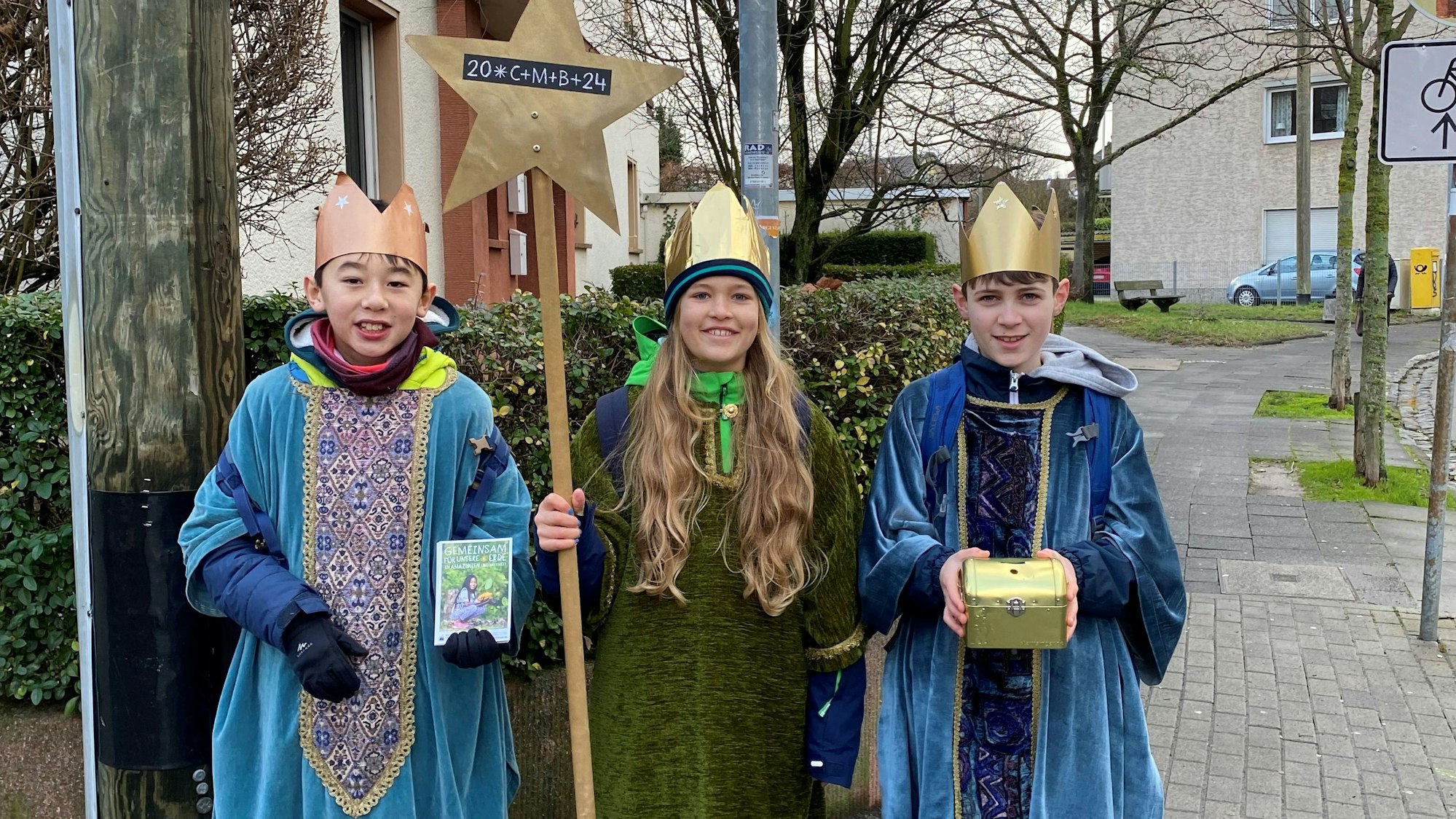 Drei Sternsinger stehen in Rheinbach auf dem Bürgersteig und halten ihre Spendendose, den Stern und Flyer mit Informationen hoch.