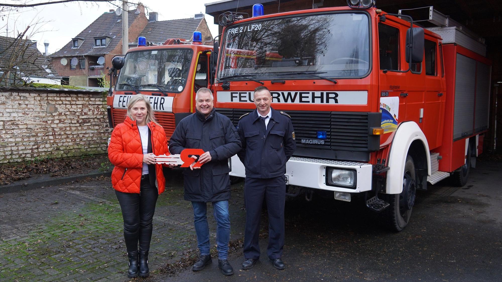 Drei Personen stehen vor zwei Feuerwehrfahrzeugen.