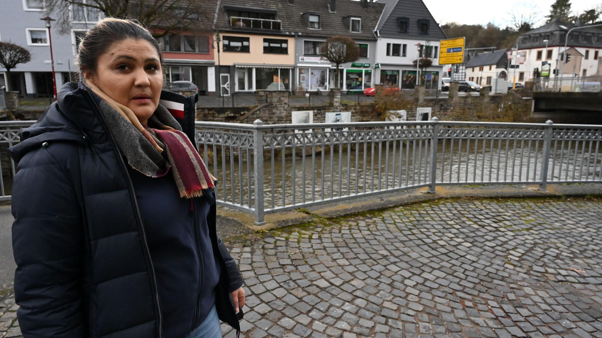 Valbone Venticinque steht am Ufer der Olef in Gemünd. Im Hintergrund sind die Kreuzung und das Geländer zu sehen, an dem sie das Auto ihrer Eltern am Morgen nach der Flut fand.