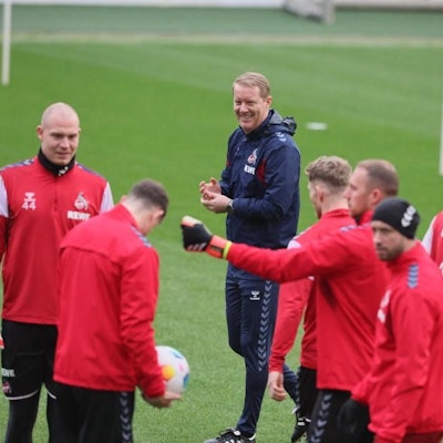Timo Schultz steht mit Spielern des 1. FC Köln auf einem Rasen.