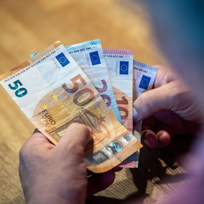 Ein älterer Mann sitzt an einem Tisch und einen 50-Euro-Schein, einen 20- und zwei 10-Euro-Scheine in der Hand.