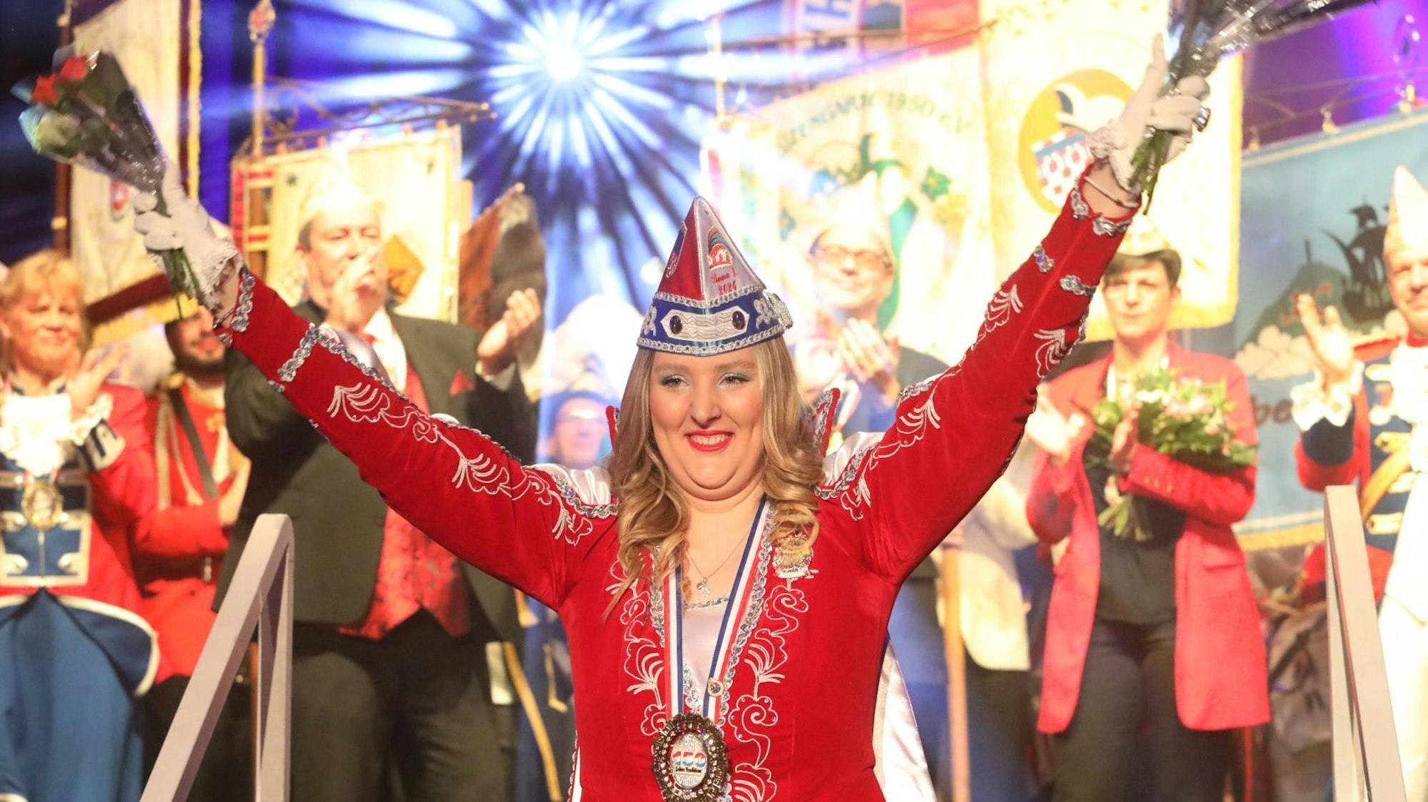 Eine junge Frau in rotem Ornat hat die Arme nach oben gerissen, im Hintergrund applaudieren auf der Bühne einige Jecken..