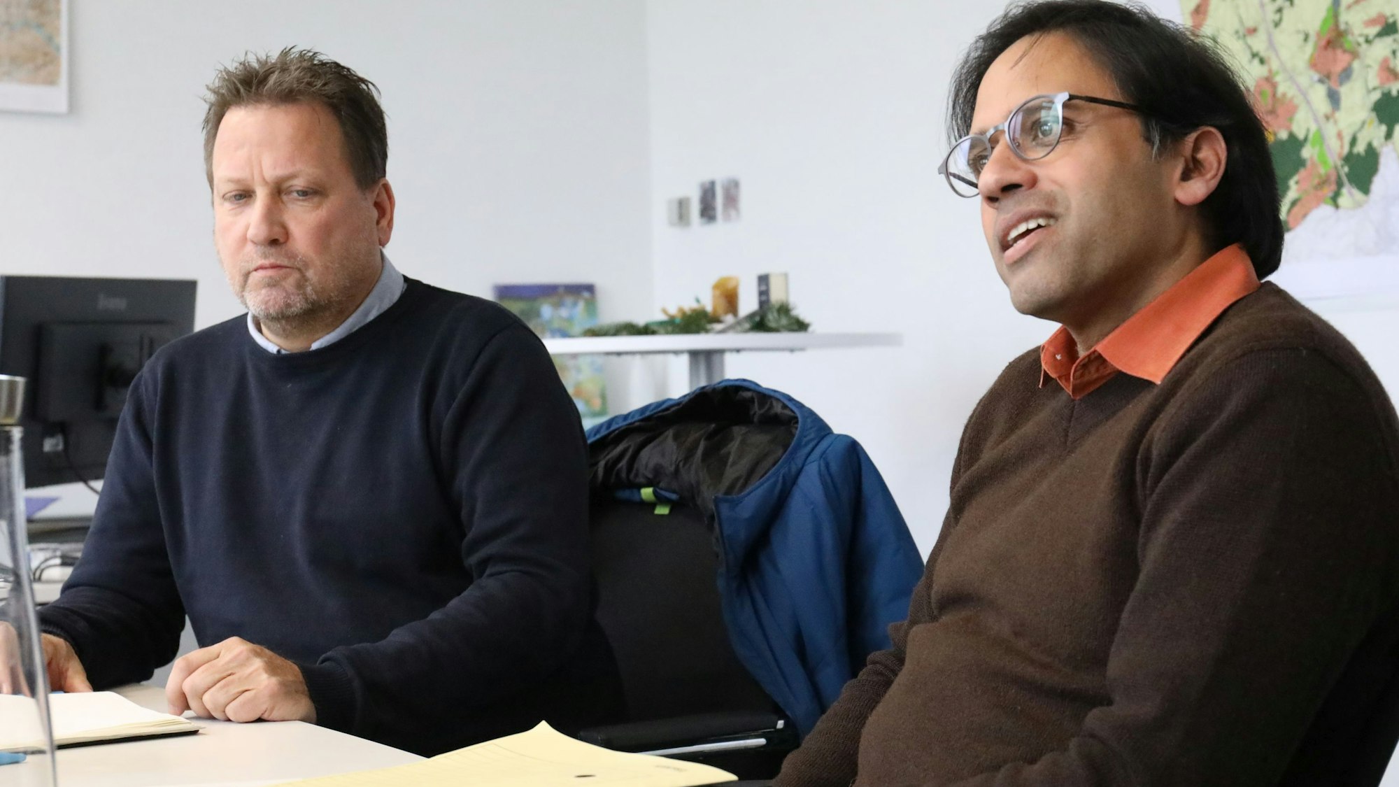 Zwei Männer sitzen an einem Tisch, auf dem Aktenmappen liegen. Im Hintergrund hängt eine mehrfarbige Stadtkarte an der Wand.