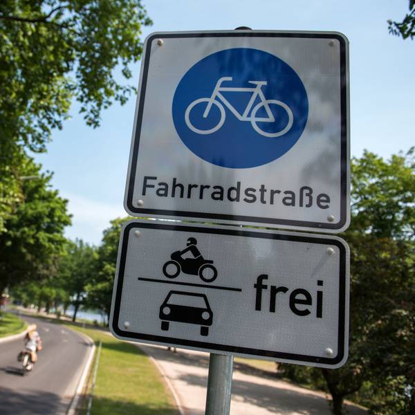 Ein Schild Fahrradstraße mit dem Zusatz Motorräder und Autos frei hängt an einer Fahrradstraße, auf der eine Radfahrerin unterwegs ist.