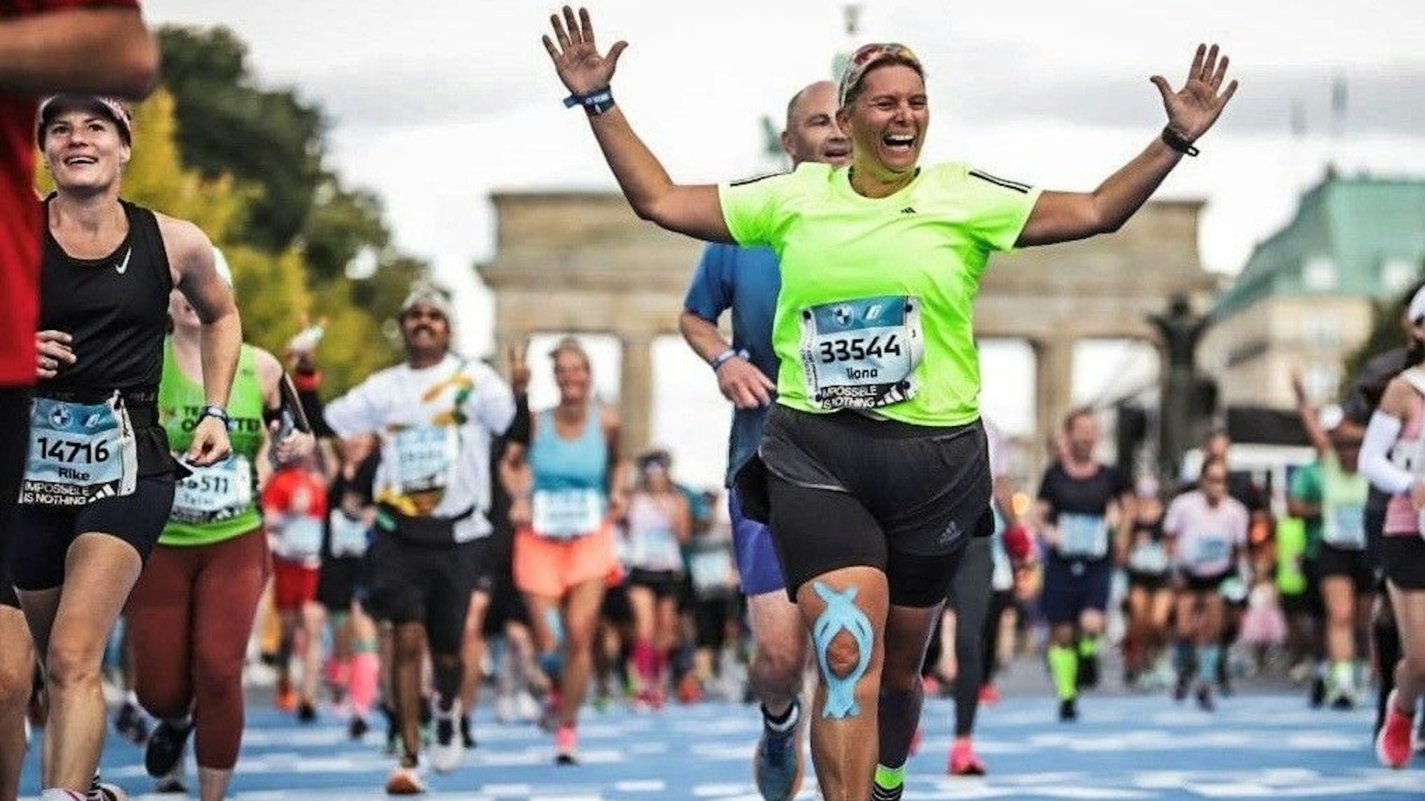 Die Läuferin Ilona Schaffrath (grünes T-Shirt) läuft beim Berlin Marathon jubelnd über die Ziellinie. Im Hintergrund ist das Brandenburger Tor zu erkennen.