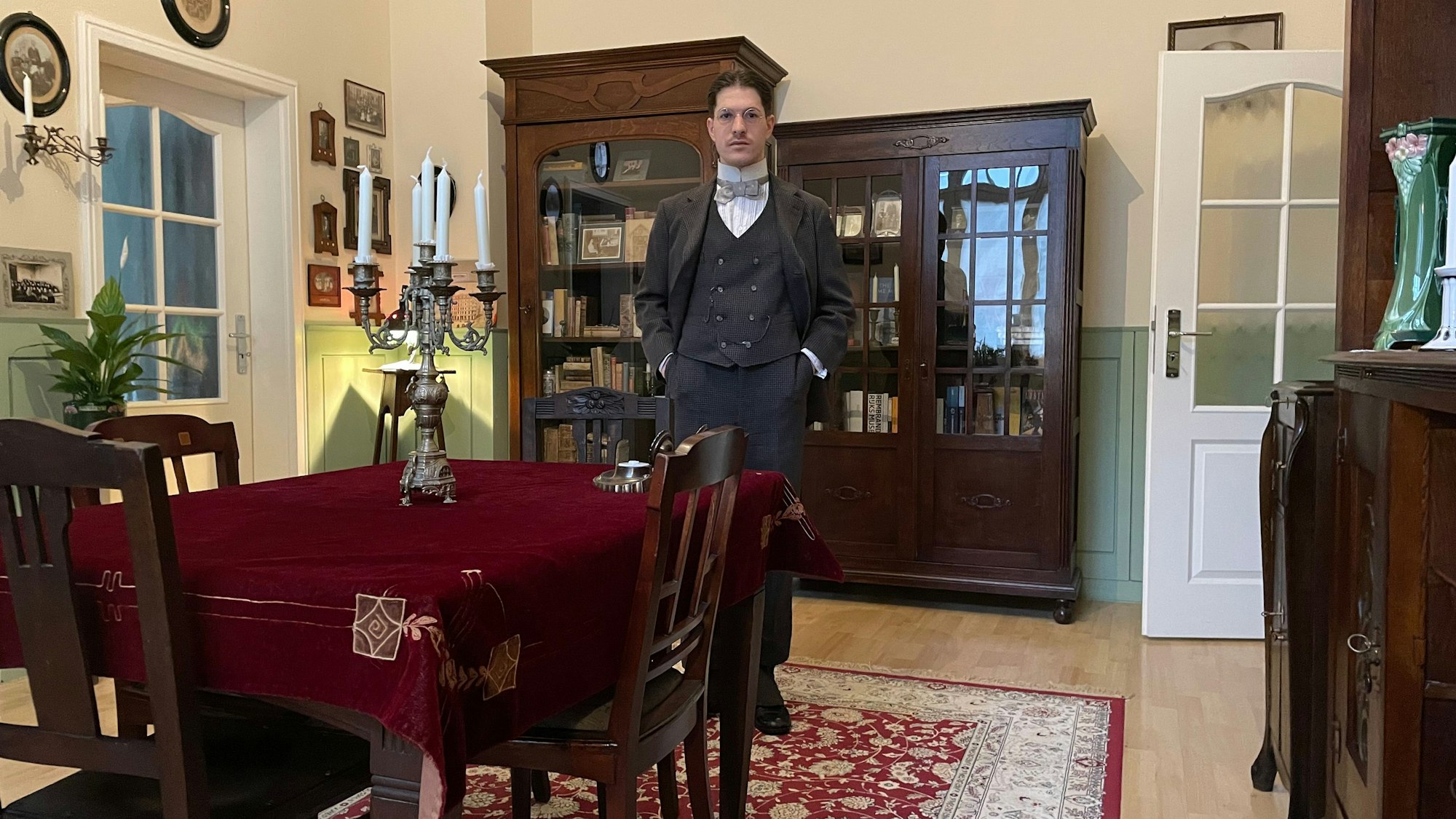 Ein Mann im altmodischen Anzug und Hemd mit Fliege steht in einem altmodisch eingerichteten Wohnzimmer.