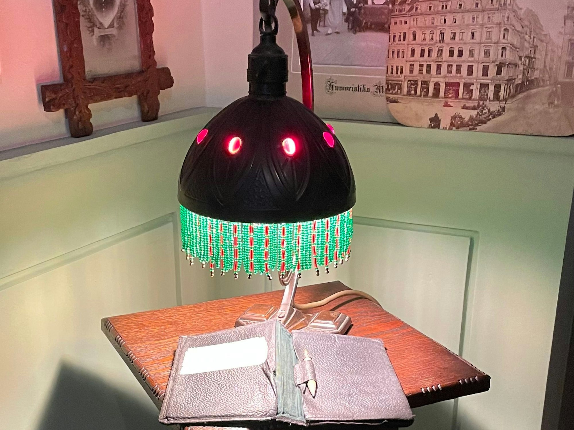 Auf einem hochbeinigen Tischchen steht eine Tischleuchte mit Fransen, an der Wand hängen alte Schwarz-Weiß-Fotos.