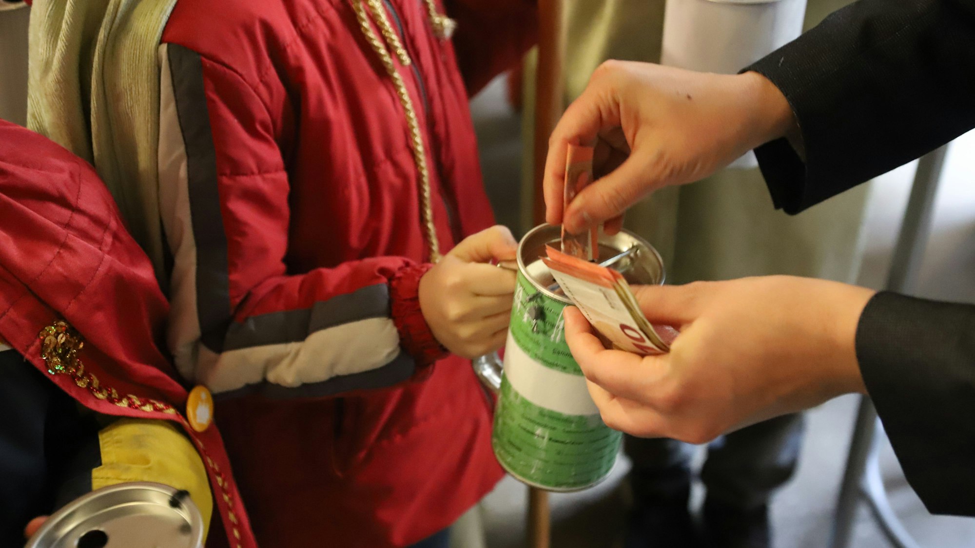 Eine Kinderhand hält eine Sammelbüchse, in die jemand Zehn-Euro-Scheine steckt.