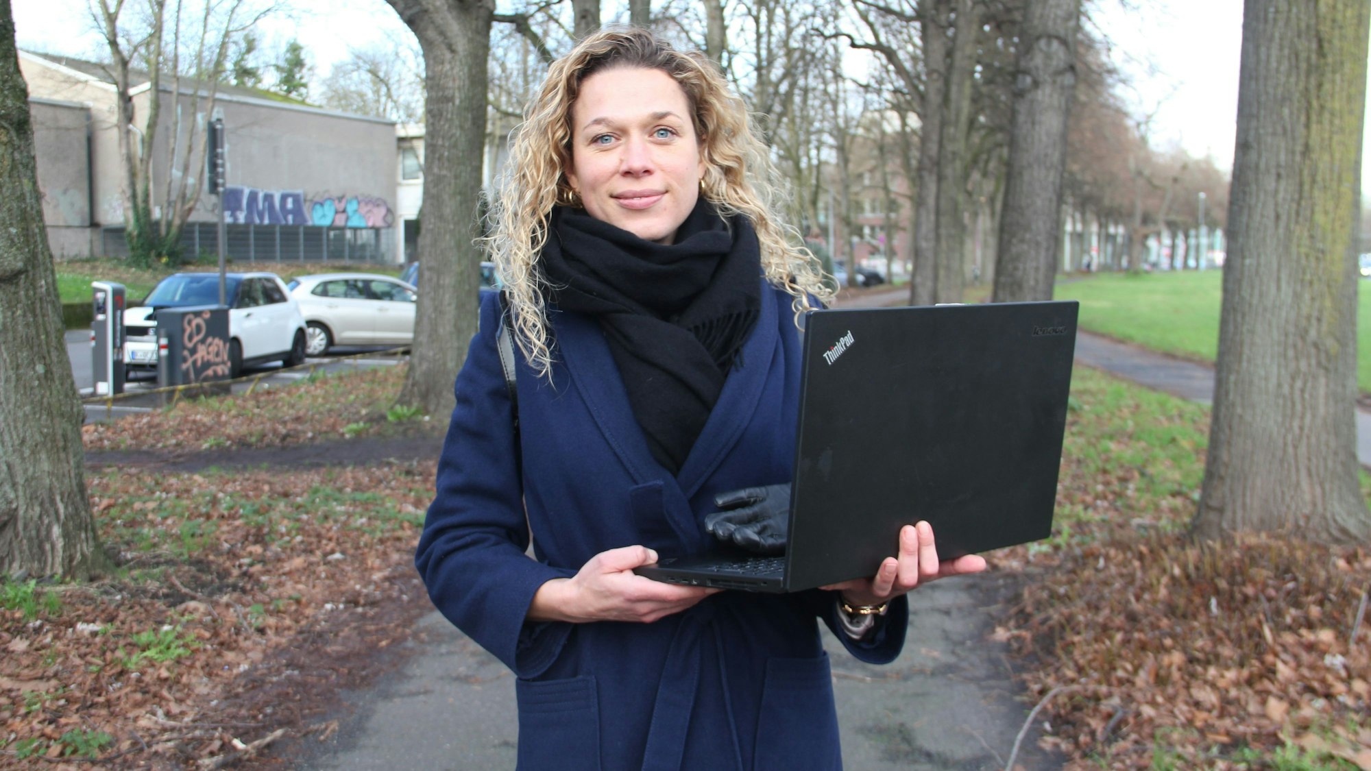 Eine blonde junge Frau steht mit einem Laptop in der Hand auf einem Spazierweg zwischen hohen Bäumen.