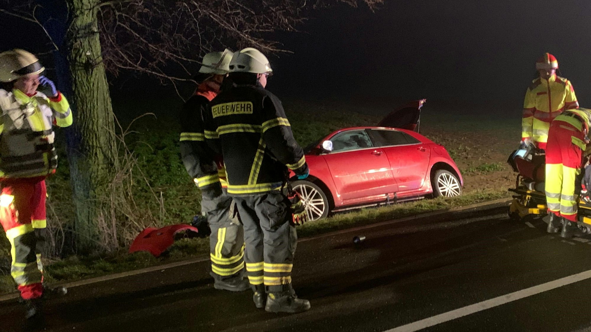 Ein rotes Auto liegt in einem Straßengraben. Davor stehen Einsatzkräfte des Rettungsdienstes und der Feuerwehr.