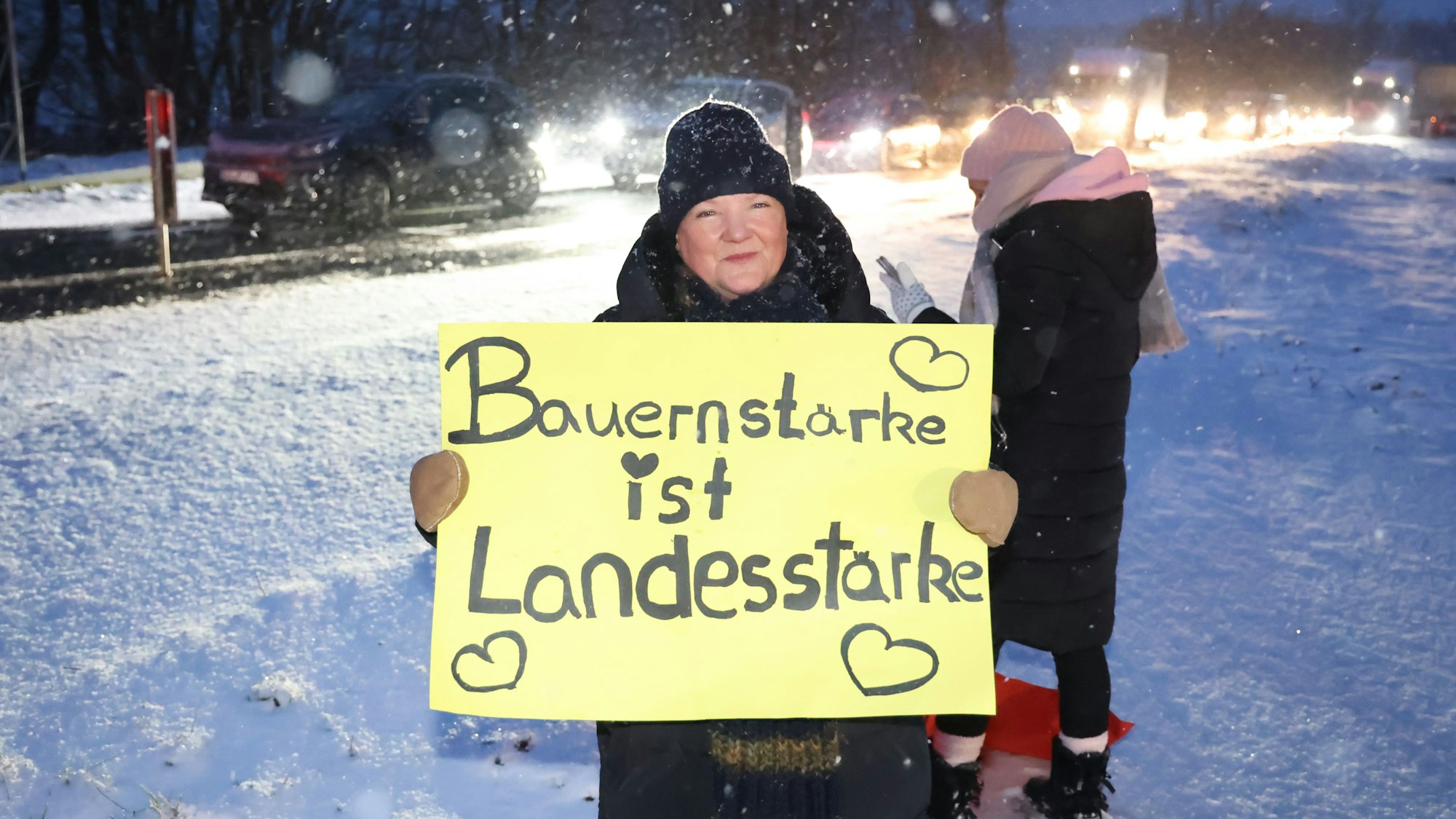 Eine Frau steht im Schneegestöber an der B51 bei Blankenheim und bekundet mit einem Transparent ihre Solidarität mit den Landwirten. Deren Demonstrationszug ist im Hintergrund zu sehen.