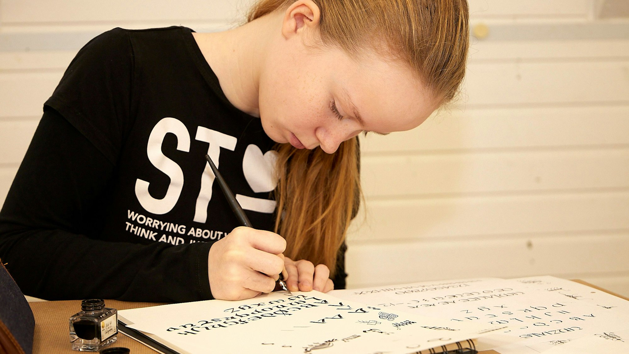 Ein Mädchen sitzt an einem Tisch und zeichnet auf einem weißen Blatt Papier kunstvolle Buchstaben.