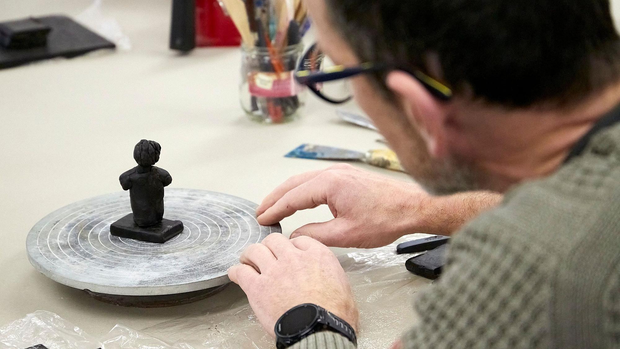 Eine kleine Skulptur aus Wachs steht auf einer drehbaren Scheibe. Ein Teilnehmer der Kunstveranstaltung schaut die Figur an, die er bearbeitet.