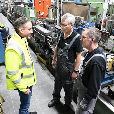 Schoeller-Geschäftsführer Alexander Mertens trägt eine gelbe Jacke. Er steht einer der Werkhallen in Hellenthal und unterhält sich mit zwei Mitarbeitern.