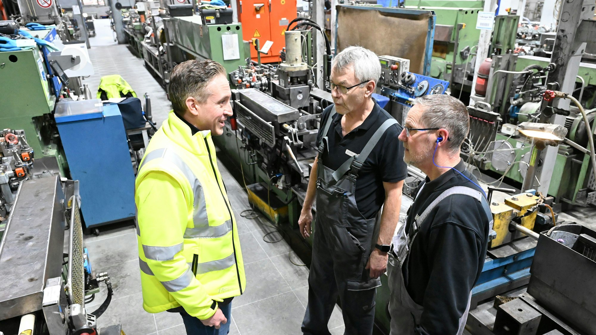 Schoeller-Geschäftsführer Alexander Mertens trägt eine gelbe Jacke. Er steht einer der Werkhallen in Hellenthal und unterhält sich mit zwei Mitarbeitern.