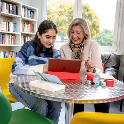 Eine Schülerin liest gemeinsam mit einer Lesementorin in der Schulbibliothek auf einem Tablet-Computer.