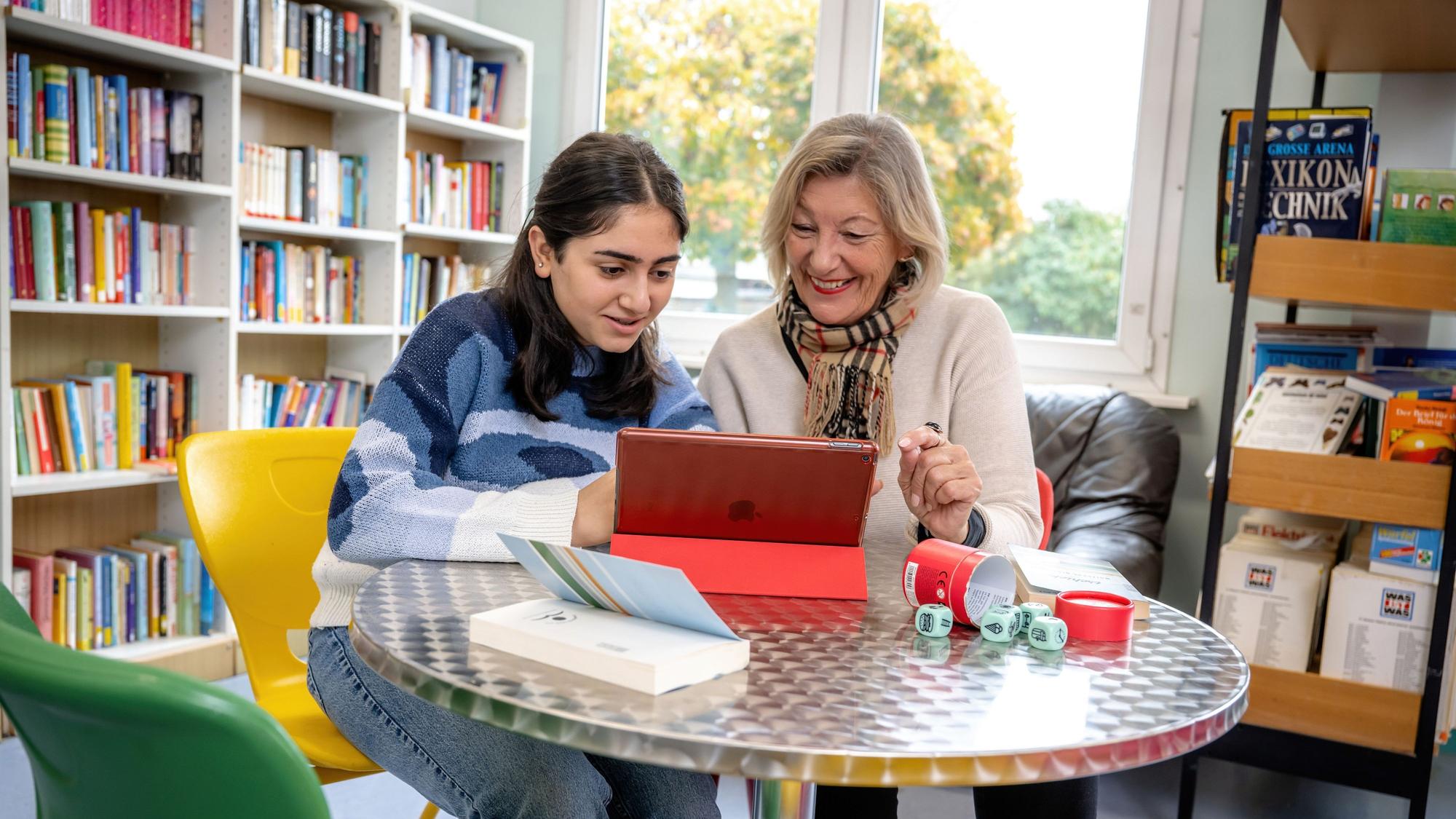 Eine Schülerin liest gemeinsam mit einer Lesementorin in der Schulbibliothek auf einem Tablet-Computer.