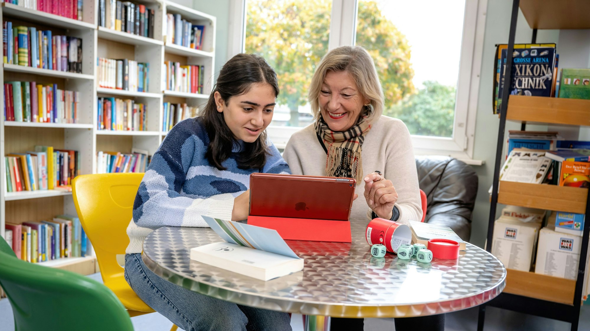 Eine Schülerin liest gemeinsam mit einer Lesementorin in der Schulbibliothek auf einem Tablet-Computer.