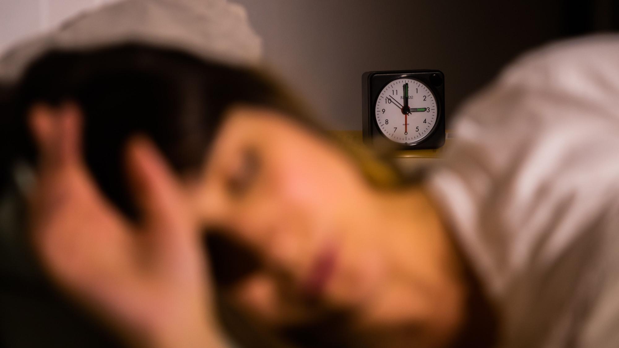 Eine Frau schläft neben einem Wecker, der auf drei Uhr zeigt.
