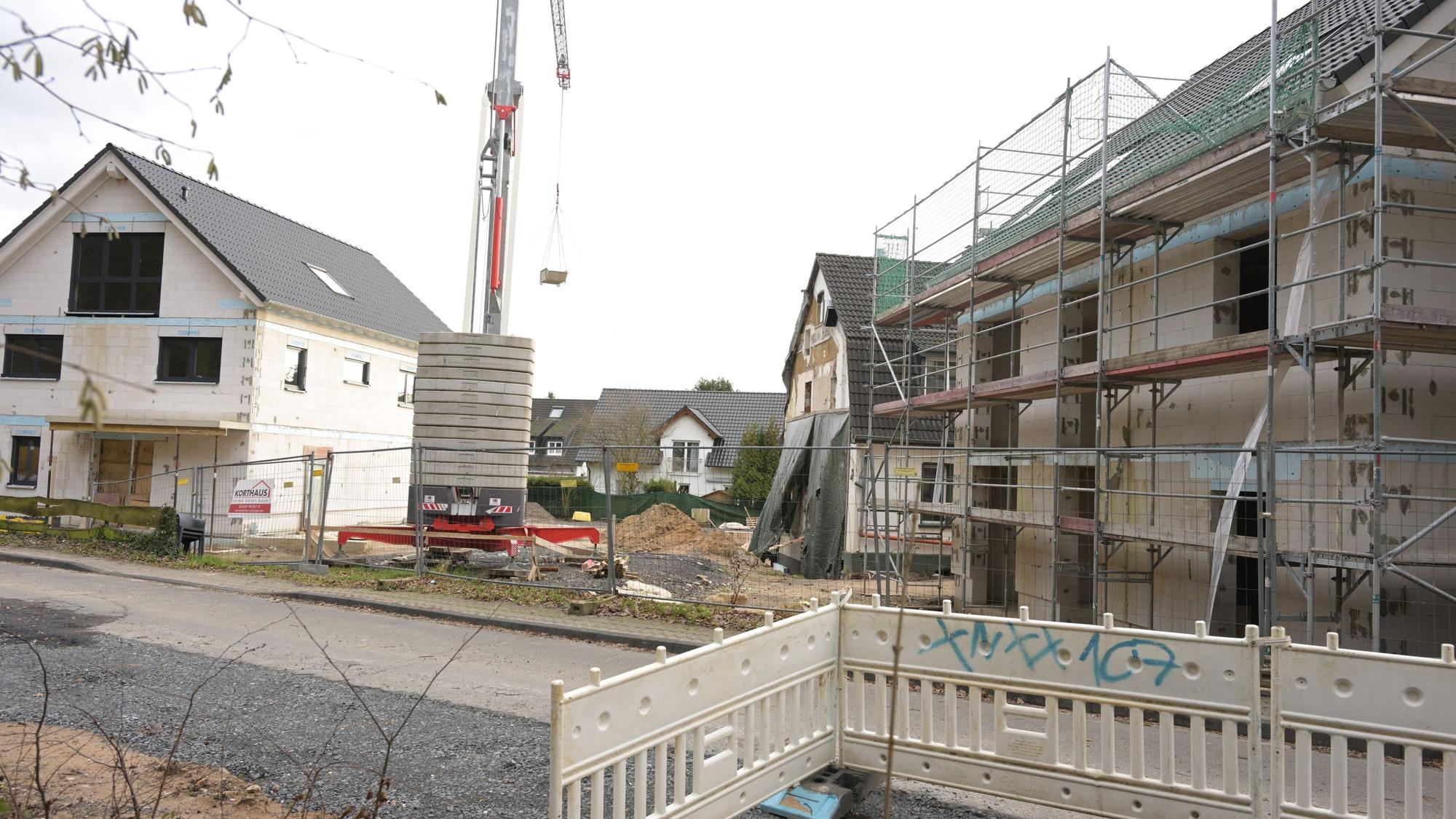 Im Vordergrund stehen Absperrungen, dahinter auf dem Baugrundstück rechts ein Rohbau mit Gerüst und links ein Einfamilienhaus, in das schon die Fenster eingesetzt sind. Dazwischen ist ein Kran platziert.