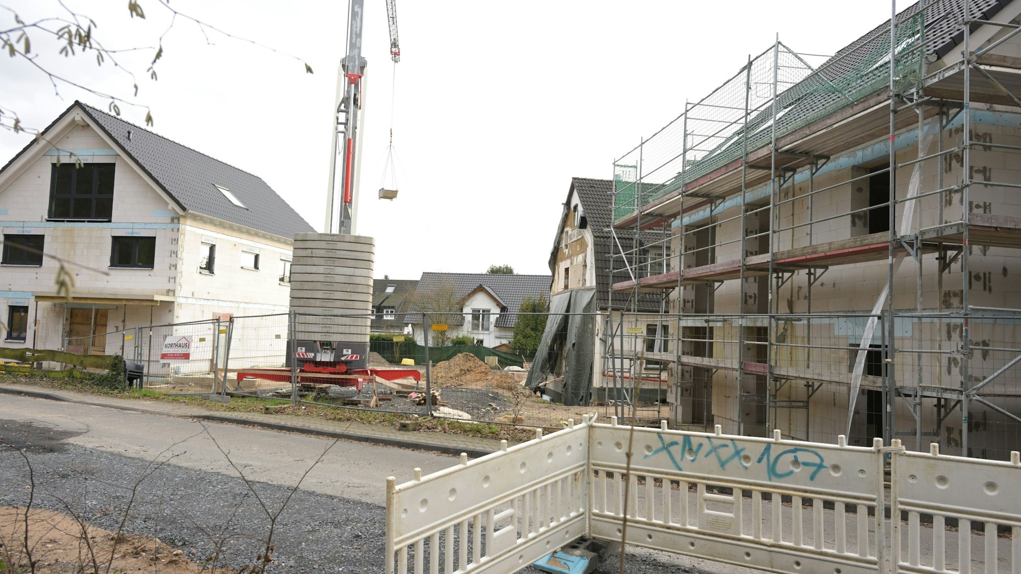 Im Vordergrund stehen Absperrungen, dahinter auf dem Baugrundstück rechts ein Rohbau mit Gerüst und links ein Einfamilienhaus, in das schon die Fenster eingesetzt sind. Dazwischen ist ein Kran platziert.