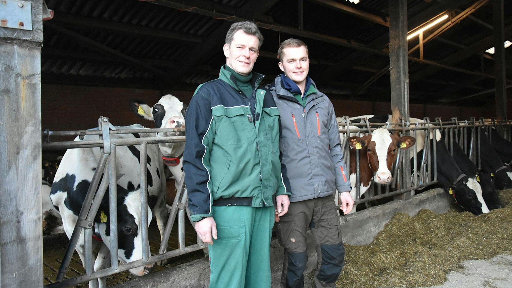 Ein Mann und sein Sohn stehen in einem Stall. Im Hintergrund sind mehrere Kühe zu sehen.