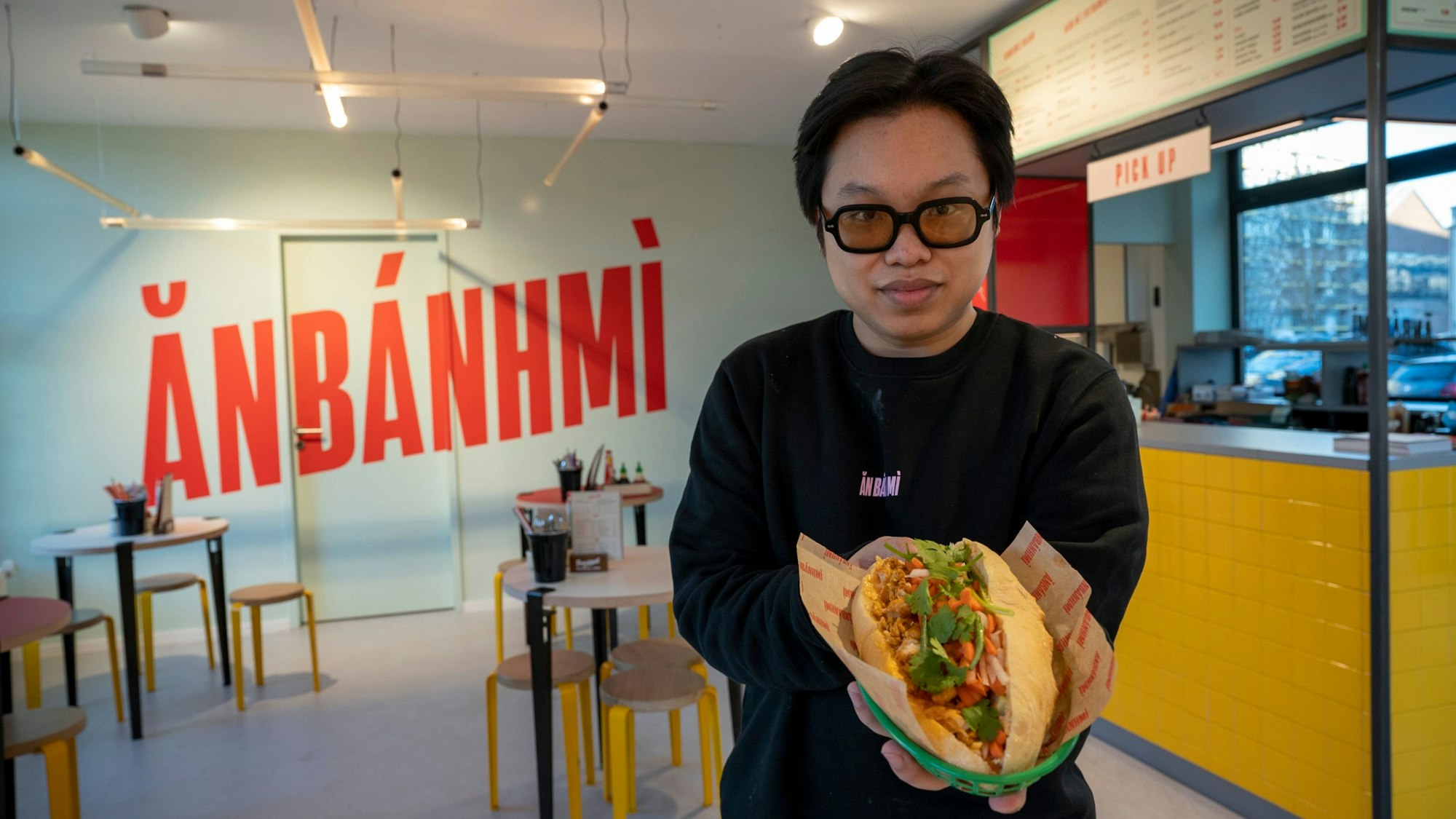 Ein Mann steht in einem Restaurant und hält ein Sandwich in die Kamera.