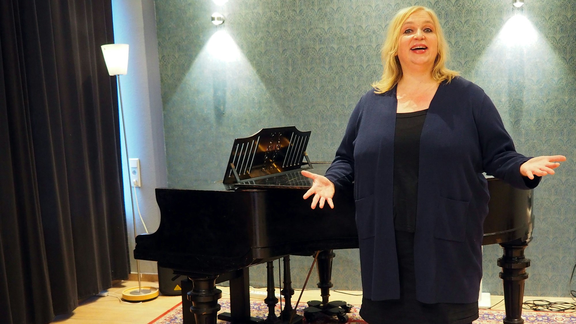 Eine blonde Frau steht mit ausgebreiteten Armen vor einem Flügel.