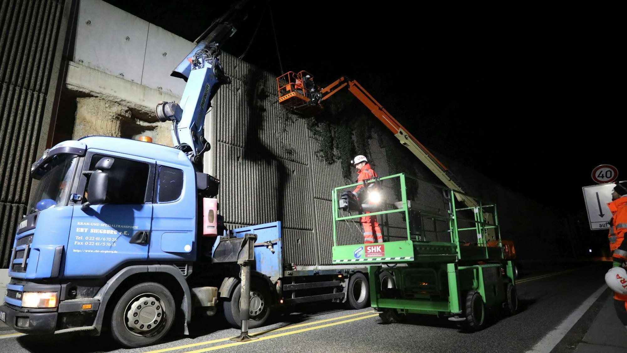 Ein blauer Lastwagen mit Anhänger und ein grüner Hubsteiger stehen nachts neben einer hohen Betonwand. Die Bundesstraße ist gesperrt.
