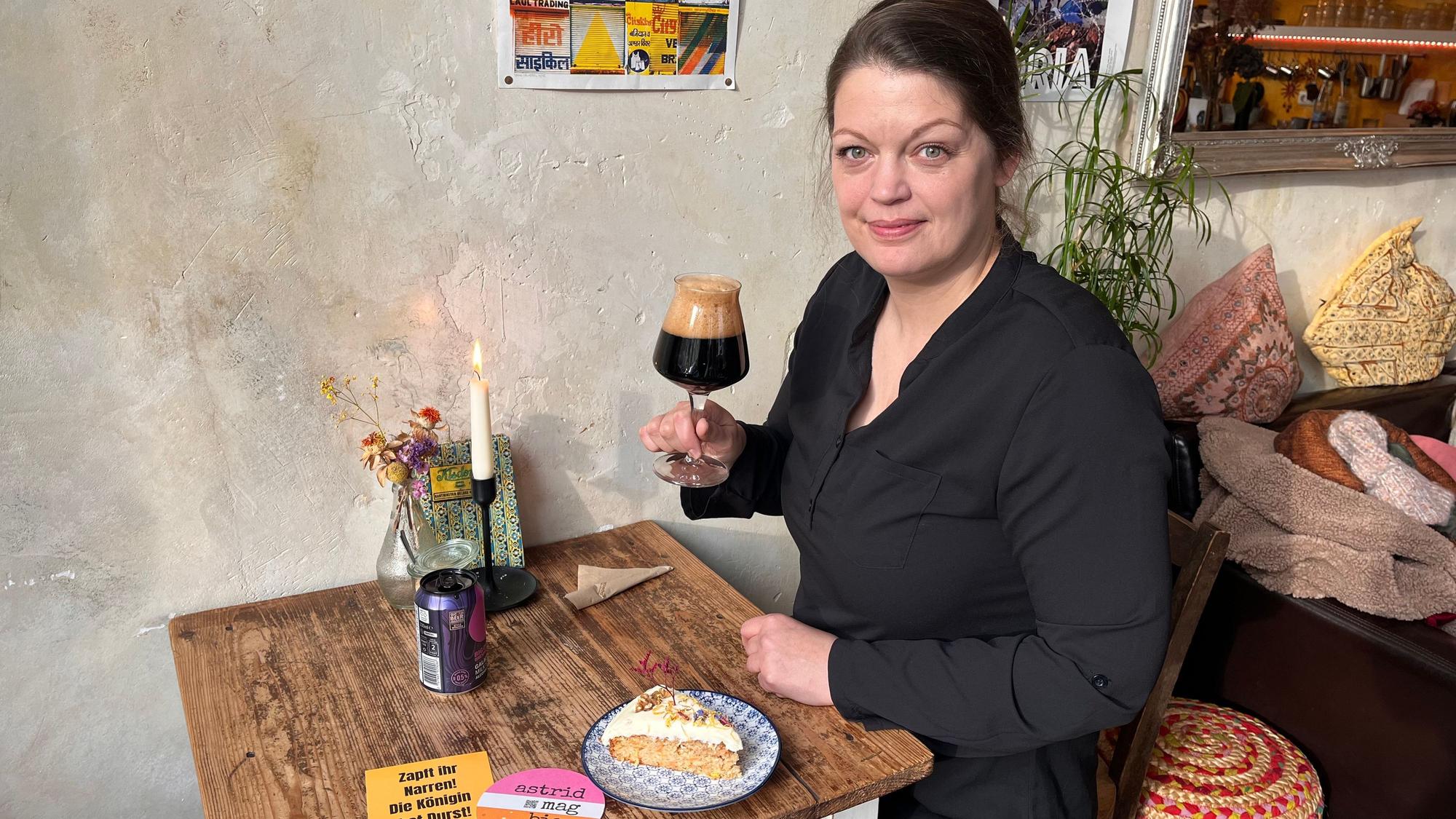 Eine Frau hat sitzt an einem Café-Tisch mit einem Glas Bier in der Hand und einem Stück Kuchen.