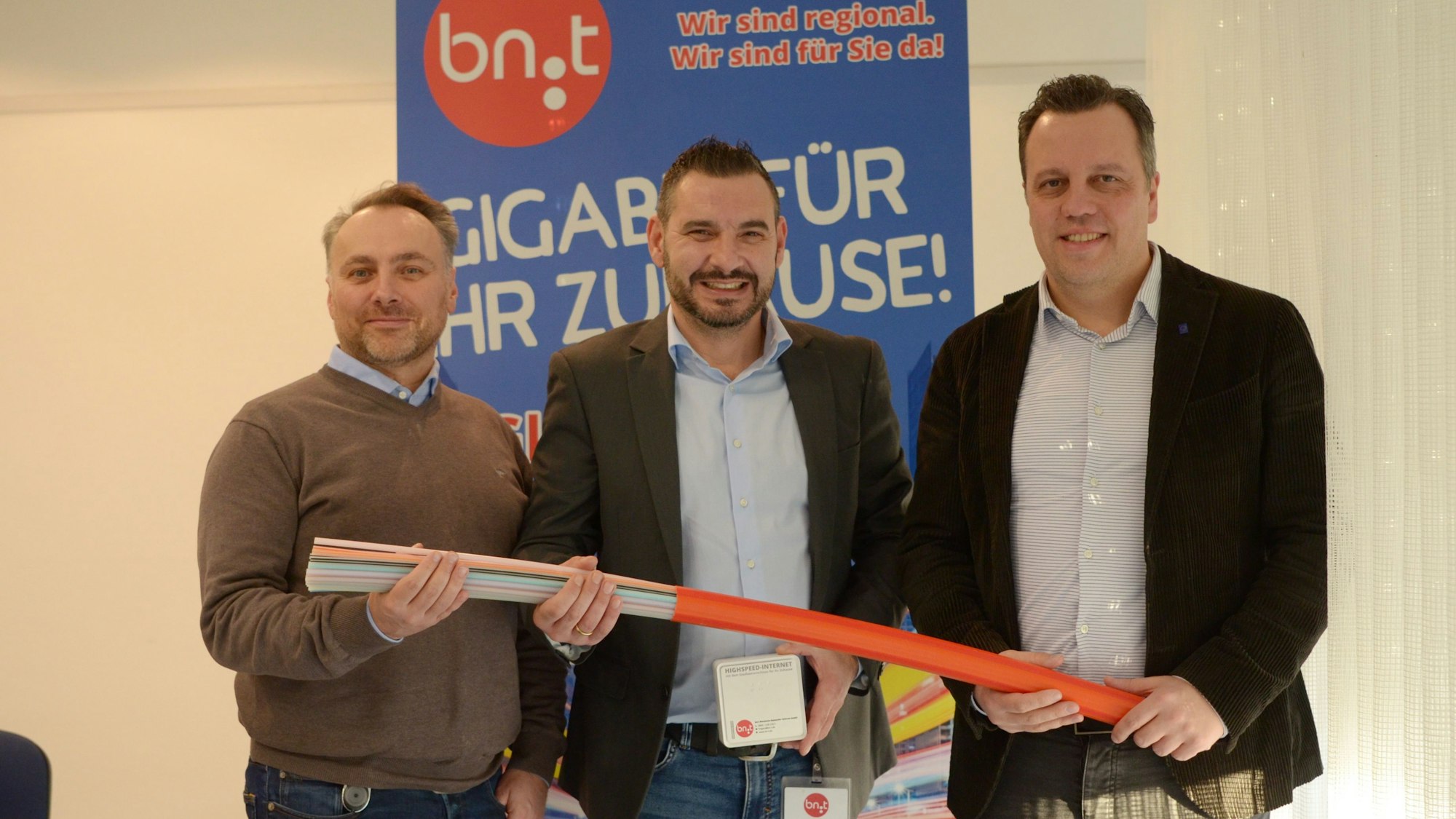 10.01.2024 Lukas Romanowski (v.l.), Nikolaos Pantios und Sacha Reichelt halten ein Glasfaserkabel in ihren Händen. Hinter ihnen ein Werbeplakat der Firma Bn:T.