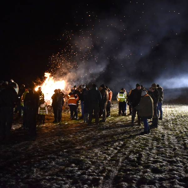 Eine Gruppe Menschen steht im Dunkeln an einem großen Feuer, rechts im Bild ist ein Traktor mit angeschaltetem Licht zu sehen.