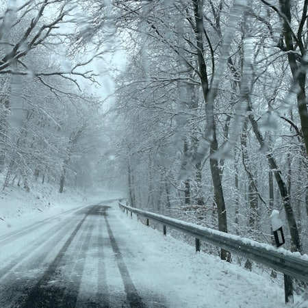 Eine eingeschneite Straße in der Eifel ist verlassen. Links und rechts der Fahrbahn liegen Schneemassen. (Archivbild)